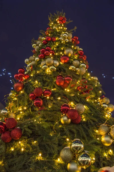 Árbol de Navidad ricamente decorado con luces de colores y globos brillantes en la noche, al aire libre, contra un cielo de invierno azul oscuro, vista inferior, vertical — Foto de Stock