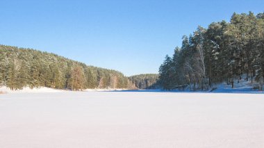 Buz ve karla kaplı donmuş bir gölün kış manzarası, kozalaklı ormanlarla çevrili, güneşli soğuk bir gün..