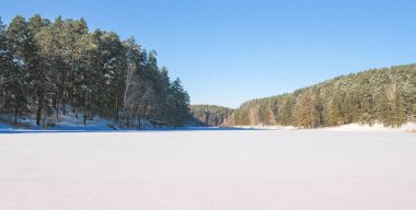 Buz ve karla kaplı donmuş bir gölün kış manzarası, kozalaklı ormanlarla çevrili, güneşli soğuk bir gün..
