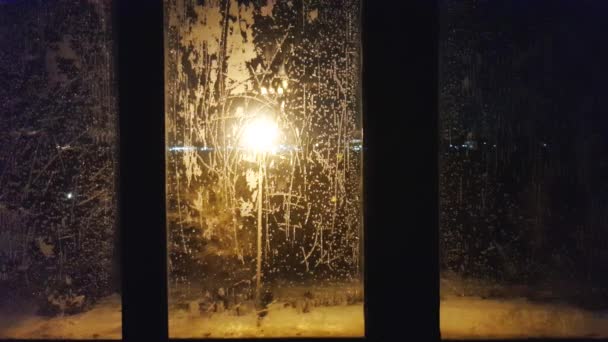 Vídeo 4k, através do vidro congelado da janela, você pode ver uma lanterna brilhando durante a noite, uma paisagem noturna da cidade com brilho e luzes no inverno, em uma noite gelada — Vídeo de Stock