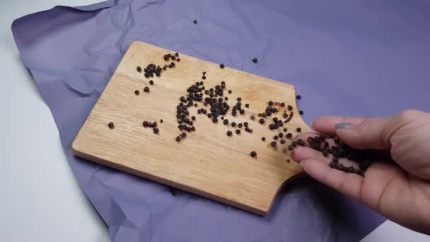 Mazăre de piper negru turnată pe o placă de tăiere din lemn, întinsă pe hârtie sfărâmată, o mână femeiască toarnă condimente — Videoclip de stoc