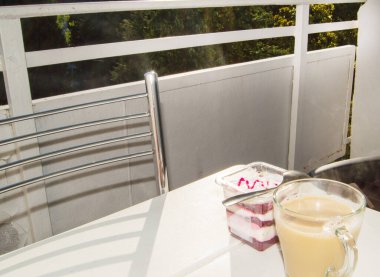 Evde ya da otelde balkonun terasında kahvaltı, çilek reçelli kremalı yoğurt ve latte kahvesi. Seçici odaklanma. Diyet kahvaltı, masa manzarası, güneş ışığı ve gölgeler..