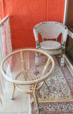 Balkonda konforlu bir oturma odası. Rat hasır bir sandalye, şeffaf bir cam masa ve bir halı, dikey bir fotoğraf..