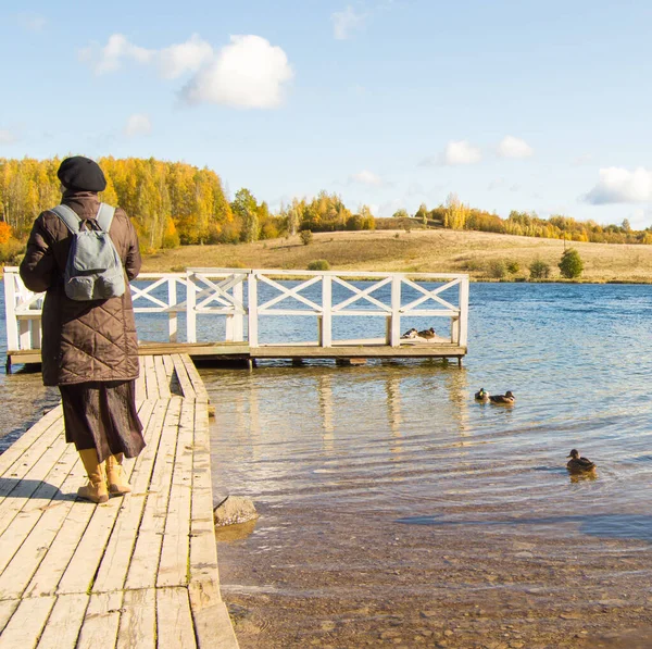 A woman in a jacket and boots, with a backpack, walks along a wooden pier in a natural park and admires the birds. Ducks in the water around.