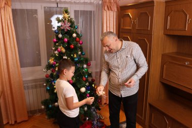 Baba ve oğul yılbaşını ya da yılbaşını kutlamak için Noel ağacının yanında havai fişek yakıyor..