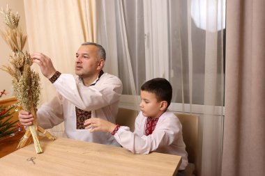 İşlemeli gömlekler içindeki baba ve oğul, zenginliğin sembolü olan buğday saplarından yapılmış didukh 'u inceleyin..