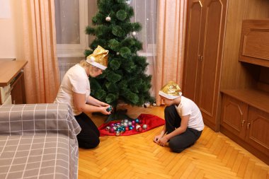 Anne ve çocuk odada bir Noel ağacı süsler, altın şapkalar takar, rahat bir şenlik ortamı hazırlarlar..