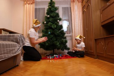 Anne ve çocuk odada bir Noel ağacı süsler, altın şapkalar takar, rahat bir şenlik ortamı hazırlarlar..