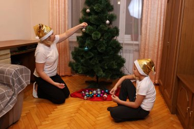 Anne ve çocuk odada bir Noel ağacı süsler, altın şapkalar takar, rahat bir şenlik ortamı hazırlarlar..