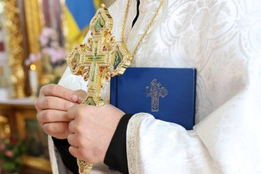 Açık renk cüppeli bir rahip elinde süslü bir haç ve kilisedeki inanç ve maneviyatı simgeleyen mavi bir kitap tutuyor..