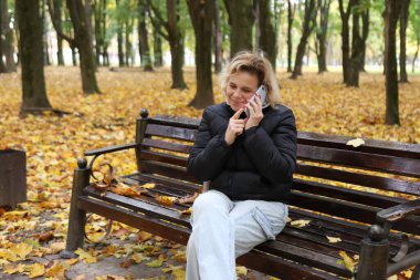 Bir kadın, güzel bir sonbahar gününde parkta bir bankta otururken telefonla konuşuyor..
