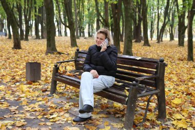 Bir kadın, güzel bir sonbahar gününde parkta bir bankta otururken telefonla konuşuyor..