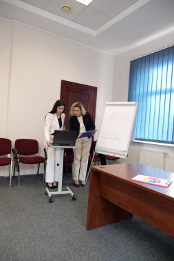 Ofisteki iki kadın bir çizelgenin yanında duruyor, belgeleri inceliyor ve bir iş sunumu sırasında verileri tartışıyorlar..