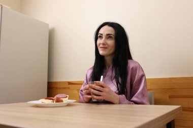 Bir kadın mutfakta masada otururken kahve ve sandviçle kahvaltı yapıyor..