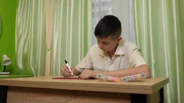 Bir çocuk parlak bir odada masasında kalemlerle çizim yapıyor. Etrafı rahat bir ev atmosferiyle çevriliyken sanat eserlerine odaklanıyor..