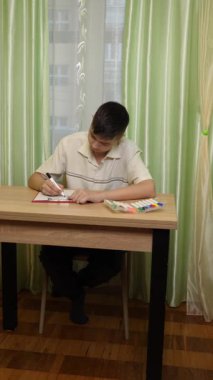 Bir çocuk parlak bir odada masasında kalemlerle çizim yapıyor. Etrafı rahat bir ev atmosferiyle çevriliyken sanat eserlerine odaklanıyor. Dikey video