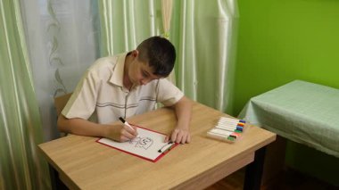 Bir çocuk parlak bir odada masasında kalemlerle çizim yapıyor. Etrafı rahat bir ev atmosferiyle çevriliyken sanat eserlerine odaklanıyor..