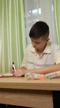 Bir çocuk parlak bir odada masasında kalemlerle çizim yapıyor. Etrafı rahat bir ev atmosferiyle çevriliyken sanat eserlerine odaklanıyor. Dikey video