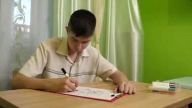 Bir çocuk parlak bir odada masasında kalemlerle çizim yapıyor. Etrafı rahat bir ev atmosferiyle çevriliyken sanat eserlerine odaklanıyor..