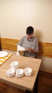 Bir adam mutfakta masada otururken bulaşıkları yıkayıp parlatıyor..