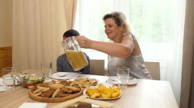 Bir kadın masada otururken oğlunun bardağına meyve suyu döker..