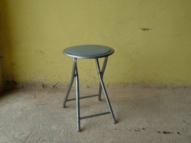 Gri metal katlama taburesi beton bir zeminde duruyor sarı bir duvarın yanında..