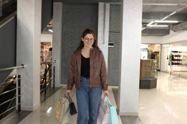 Alışveriş çantalı genç bir kadın alışveriş merkezinde yürüyor, başarılı alışveriş deneyimlerinden sonra gülümsüyor.