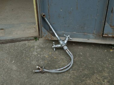 Metal su musluğu, esnek hortumu eski metal kapıların yanında betonun üzerinde duruyor..