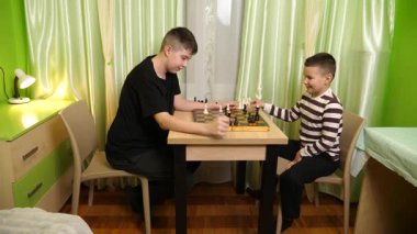 İki kardeş sıcak yeşil bir odada, masada satranç oynuyorlar. Düşünceli bir şekilde hareketlerin tadını çıkarıyorlar ve sessiz etkileşimlerin tadını çıkarıyorlar..