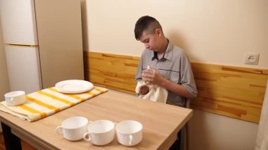 Genç bir çocuk, bir evde mutfakta bulaşıkları kurutuyor ve temizliyor. Kendi başına günlük ev işlerini yapıyor..
