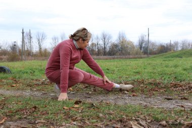 Kadın yerde otururken bacağını uzatır, sonbahar doğasında sakin esneklik egzersizi yapar..