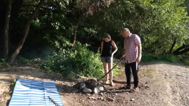 Bir adam kamp ateşine doğru eğiliyor ve bir kadın dışarıda uzun bir yemek pişirme işlemi sırasında yanında duruyor.