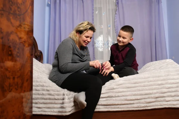 Anne yatakta oturup oğlunun tırnaklarını törpülüyor, evde bakım ve aile bağlarını kuruyor.