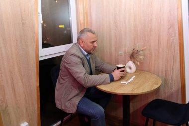 Pencerenin yanındaki kafede oturan, elinde içki tutan ve düşünceli bir şekilde masaya bakan bir adam..