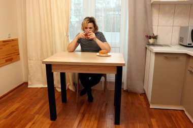 Mutfak masasında oturan, çörekle çay içen, pencerenin yanındaki fincana bakan bir kadın..