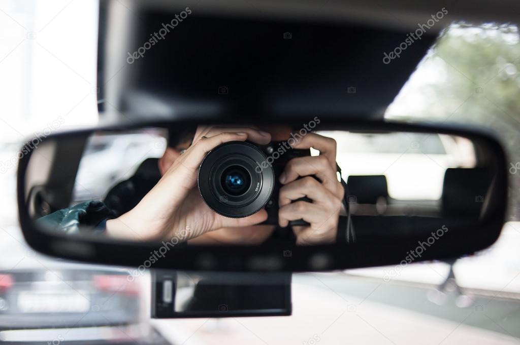 man holding a camera sits on driver's seat and Looks in the rear-view mirror
