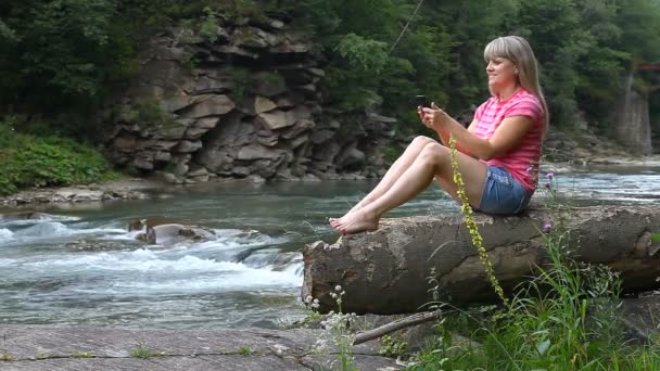 Mladá dívka sedí u řeky a samotné fotografování na smartphone. — Stock video