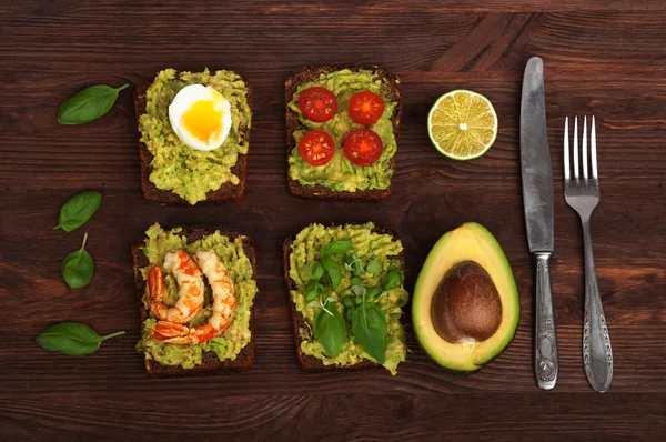 Sandwiches mit Guacamole mit verschiedenen Füllungen wie Ei, Kirschtomaten, Garnelen und Basilikumblättern auf braunem Holzgrund. in der Nähe von Besteck und der Hälfte der Avocado. gesunde Snacks hausgemacht. — Stockfoto