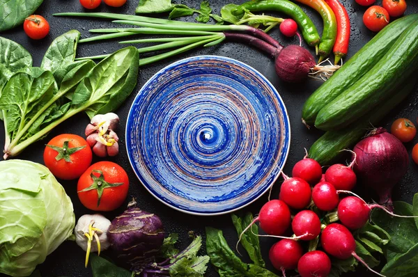The blue plate surrounded by vegetables and herbs, such as cherry tomatoes, radishes, green onions, spinach, garlic and hot peppers. Vegetable background. Vegan concept