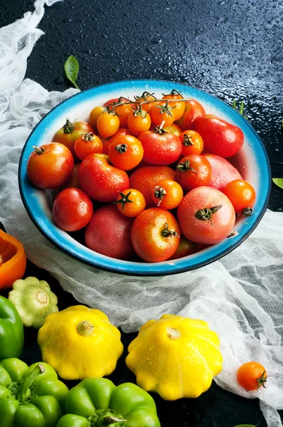 Teller mit Bio-Tomaten in verschiedenen Farben und Größen auf schwarzem Hintergrund. in der Nähe der anderen Gemüsesorten wie Paprika und Zucchini. Veganes Konzept. pflanzlicher Hintergrund (Tapete) — Stockfoto