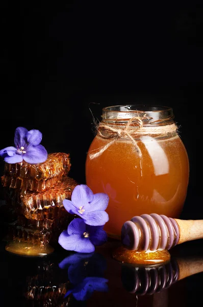 Miel en un frasco de vidrio sobre un fondo negro. Cerca de flores púrpuras y panal. Producto natural de la apicultura . — Foto de Stock