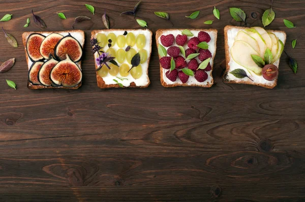 Fruit sandwiches with soft cheese and various toppings, such as figs, raspberries, slices of pear, green grapes and basil leaves. Brown wood background. Place for writing text or recipe. Ready homemade healthy snacks