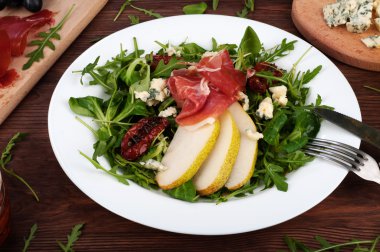 The concept of Italian food. Salad with arugula, pear slices, dried tomatoes, ham and blue cheese on dark board.