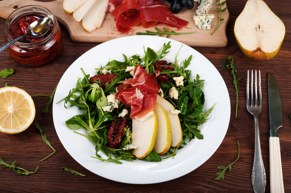 The concept of Italian food. Salad with arugula, pear slices, dried tomatoes, ham and blue cheese on dark board.