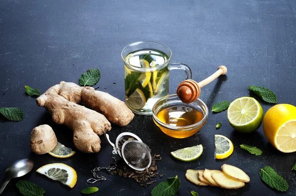 Ginger tea with lemon, lime and mint on black background. Beside the ingredients for tea, the same as the mint leaves, chopped ginger root, slices of lemon and lime. The concept of treatment of colds folk methods. — Stock Photo, Image