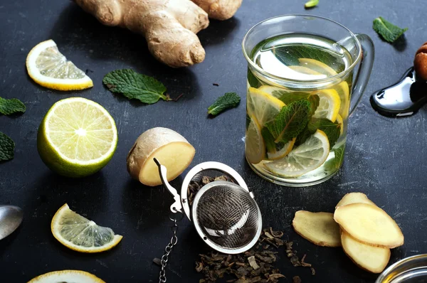 Té de jengibre con limón, lima y menta sobre fondo negro. Al lado de los ingredientes para el té, lo mismo que las hojas de menta, raíz de jengibre picada, rodajas de limón y lima. El concepto de tratamiento de los resfriados métodos populares . — Foto de Stock