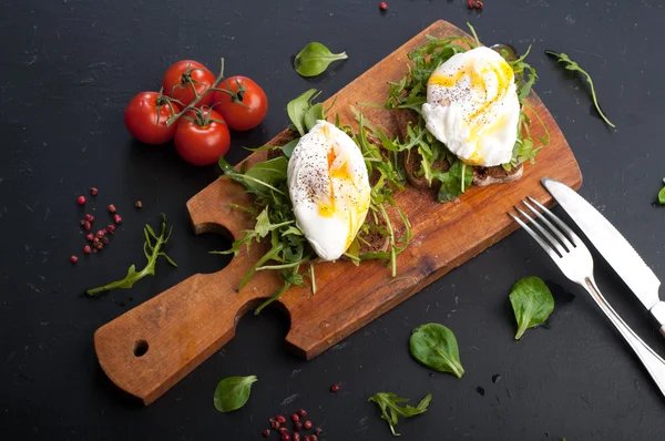 Sandwiches with arugula and poached egg on a wooden board. Near tomatoes and spices. The concept of useful home-made snacks — Stock Fotó