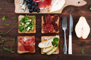 Sandwiches with a variety of toppings such as arugula, blue cheese, dried tomatoes, slices of pear, ham on a dark wooden board. Italian food concept