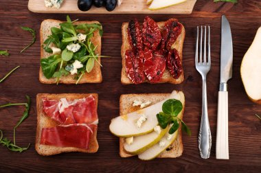 Sandwiches with a variety of toppings such as arugula, blue cheese, dried tomatoes, slices of pear, ham on a dark wooden board. Italian food concept