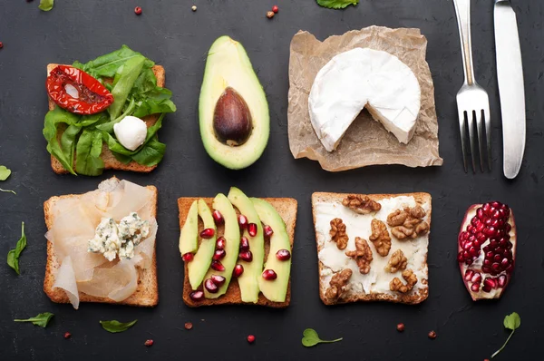 Broodjes met een verscheidenheid van toppings zoals rucola, gedroogde tomaten, zachte kaas, blauwe kaas, noten en avocado's op een donkere achtergrond. Het concept van gezonde vegetarische voeding. — Stockfoto